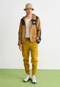 Braune und schwarze Jacke mit Kapuze, weißes Hemd, gelbe Jogginghose und beiger Bucket Hat. Die Schuhe sind weiß mit schwarzen Akzenten.