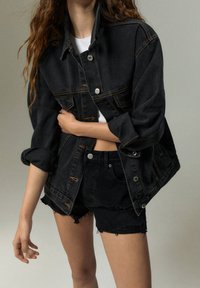 Donna che indossa una giacca di jeans nera con maniche rimboccate e pantaloni shorts in denim nero sfibrati abbinati su un top bianco a maniche corte, contro uno sfondo semplice.