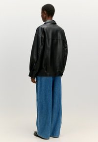 Schwarze Lederjacke mit lässiger Passform und Kragen, kombiniert mit weiten blauen Jeanshosen. Glatte Textur, minimale Details.