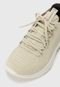 Lys beige atletiske sko med en struktureret strikoverdel, flade snørebånd og en polstret sål. Har subtile sorte detaljer på siderne.