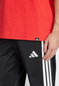 T-shirt rossa di cotone con maniche corte, abbinata a pantaloni sportivi neri con strisce bianche sui lati e un piccolo logo bianco sulla coscia.