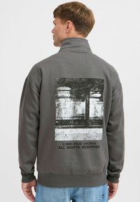 Chaqueta de pullover gris con un alto cuello, gran estampado gráfico en la parte posterior y puños acanalados. Fabricada en un tejido suave y texturizado.