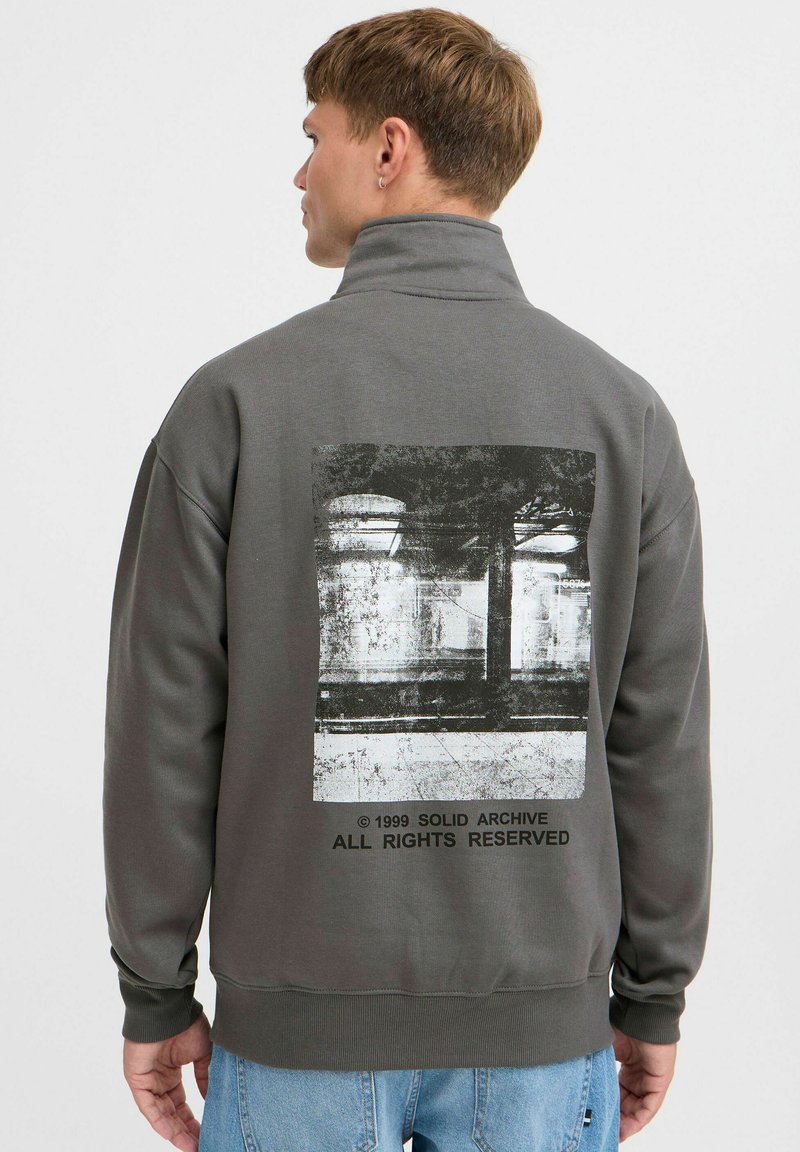 Chaqueta de pullover gris con un alto cuello, gran estampado gráfico en la parte posterior y puños acanalados. Fabricada en un tejido suave y texturizado.