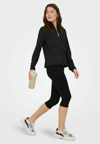 Giacca nera con zip e maniche lunghe, abbinata a leggings neri Capri. La persona tiene in mano una borraccia beige. Scarpe da ginnastica grigio chiaro e nere.