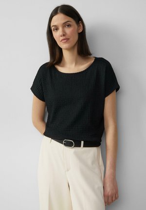 Haut noir texturé à manches courtes avec un décolleté large, associé à un pantalon crème ajusté et une ceinture noire avec des accessoires en argent.