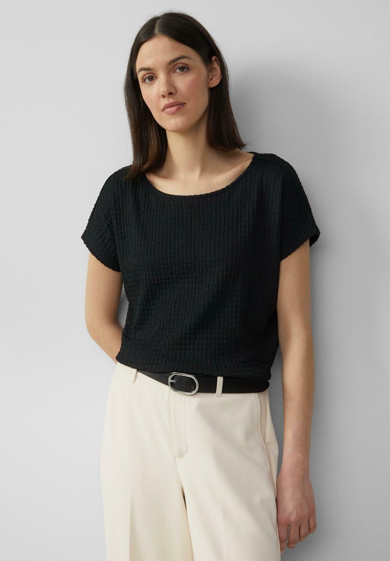 Top negro texturizado de manga corta con escote ancho, combinado con pantalones ajustados color crema y un cinturón negro con herrajes plateados.