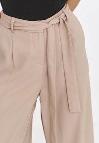 Beżowe spodnie o szerokich nogawkach z wiązanym paskiem, charakteryzujące się gładką fakturą i bocznymi kieszeniami. Materiał wydaje się lekki i delikatnie opada.