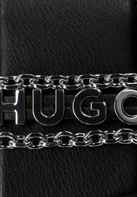 Sfondo in pelle nera con una catena argentata lucida che presenta lettere in rilievo che formano la parola "HUGO." I link della catena sono lisci e rotondi.