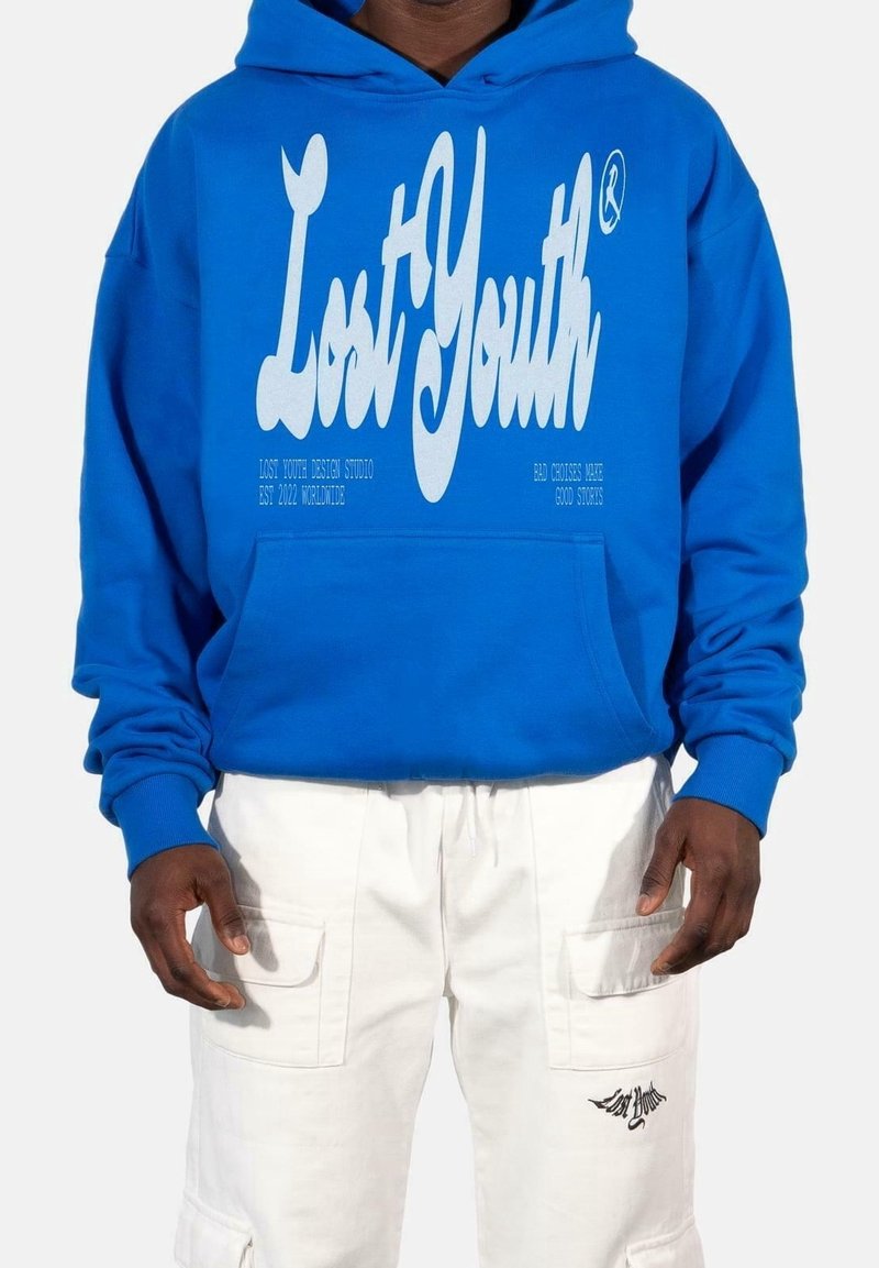 Personne portant un sweat à capuche bleu avec le texte "Lost Youth" et un pantalon cargo blanc avec des rabats de poche et un petit logo noir sur la cuisse gauche.