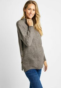 Pull en maille gris avec un col montant, des poignets côtelés, des boutons sur le côté et une coupe décontractée. Assorti avec un jean bleu, mettant en valeur la texture et la chaleur.