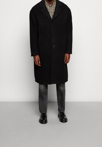 Schwarzer Wollmantel mit Reverskragen, einreihig, zwei Fronttaschen, getragen über einem karierten Hemd, kombiniert mit dunklen Jeans und Stiefeln.