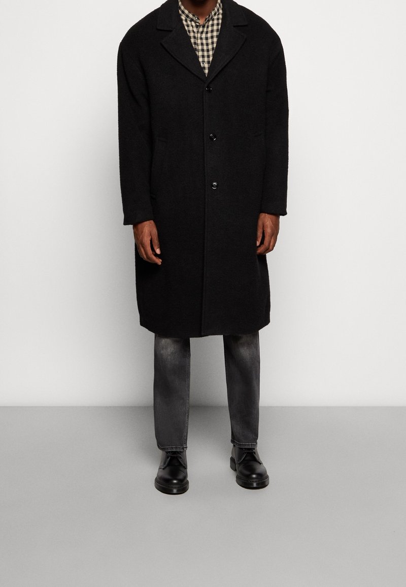 Schwarzer Wollmantel mit Reverskragen, einreihig, zwei Fronttaschen, getragen über einem karierten Hemd, kombiniert mit dunklen Jeans und Stiefeln.