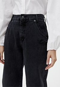 Pantalones de denim negros de cintura alta con un corte relajado, que presentan costuras visibles, bolsillos frontales y detalles desgastados.