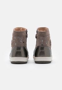 Pár dětských surových kožených kotníkových bot v hnědé a kovově šedé barvě se bočními zipy a bílou gumovou podrážkou, z pohledu zezadu.