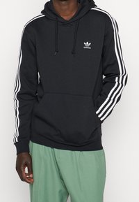 Musta hupullinen collegetakki, jossa valkoiset kolme raitaa hihoissa ja pieni valkoinen logo. Varustettu kengurun taskulla ja säädettävällä kiristysnauhalla hupussa.