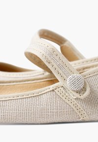 Chaussure en tissu beige avec un dessus en toile texturée, une sangle ajustable et un accent de bouton rond. Semelle plate et détails de couture simples.