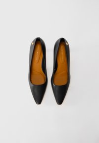 Pair of black pointed-toe Tommy Hilfiger flats with tan insoles and small gold logo embellishments near the heels on a white background.