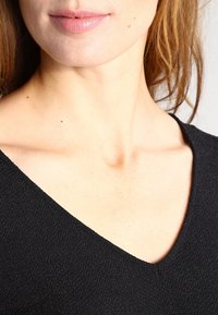 Černý top s výstřihem do V, vyrobený z texturované tkaniny, mírně přiléhavý střih, s viditelnou oblastí klíčních kostí, zaměřující se na výstřih a horní část hrudníku.