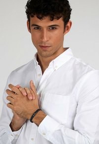 White button-up shirt with a chest pocket, featuring a textured black bracelet on the wrist. Curly hair, neutral background.