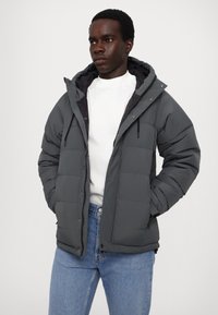 Veste rembourrée grise avec une capuche, dotée d'une fermeture éclair, de poignets élastiques et d'un design matelassé. Associée à un jean bleu et à une chemise blanche.
