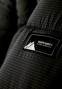 Svart puffjacka i tyg med rutmönster, med en framträdande svart och vit etikett märkt "SUPERDRY" och "Altitude Performance."