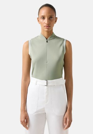 Frau trägt ärmelloses hellgrünes Zip-up-Top und hoch taillierte weiße Hose mit Gürtel, steht vor einem schlichten Hintergrund.