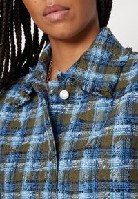 Woven Jacke in blau-grüner Karomuster, mit ausgefranstem Kragen, silbernem Knopfverschluss und strukturiertem Stoff. Nahaufnahme.