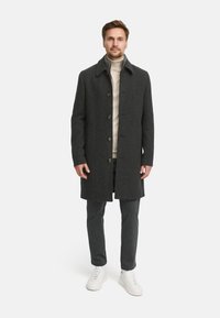 Dunkelgrauer Wollmantel mit Kragen, Knopfverschluss und Seitentaschen. Über einem hellbeigen Rollkragenpullover getragen, kombiniert mit dunklen Hosen und weißen Sneakern.