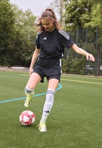 Juodas sportinis marškinėlis ir šortai su baltais įbrėžimais, balti iki kelių kojinės su juostelėmis ir geltonos spalvos batai, žaidžiant su raudonu ir baltu futbolo kamuoliu.