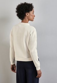 Sudadera de color beige claro con cuello redondo, puños y dobladillo acanalados. Textura suave y corte relajado, llevada sobre pantalones azul marino oscuros.