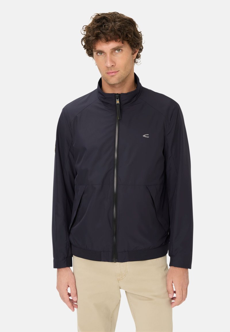 Mand iført en sort zip-up jakke med et lille logo på brystet og beige bukser, stående mod en ensartet lys baggrund.