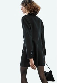 Schwarzes, maßgeschneidertes Blazerkleid mit langen Ärmeln, versehen mit Knopfdetails an den Manschetten und einem eleganten Design. Kombiniert mit einer kleinen schwarzen Handtasche.