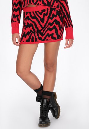 Fustă mini tricotată roșie și neagră cu model zigzag, având un brâu texturat, asociată cu cizme negre până la gleznă și șosete.