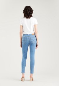 Mujer con el pelo corto y rizado de espaldas, lleva una camiseta blanca, jeans ajustados de color azul claro y sandalias de tacón cuadrado beige.