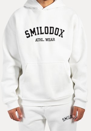 Person trägt weißen Hoodie und passende Jogginghose mit schwarzem Text "SMILODOX ATHL. WEAR" auf der Brust und "SMILODOX" auf der Hose.