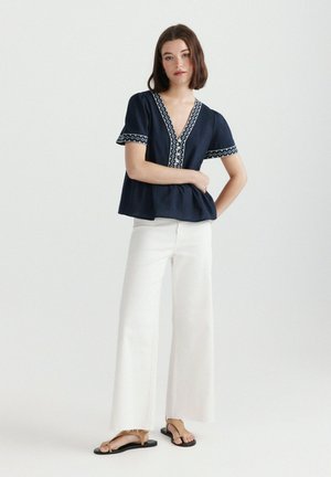 Jonge vrouw die een marineblauwe blouse met korte mouwen en witte borduursels draagt, witte wijde broeken en beige platte sandalen.