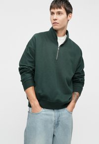 Tmavě zelená mikina s vysokým límcem a polovičním zipem. Má volné rukávy, elastické manžety a hladký textilní povrch. Nesená přes bílou košili.