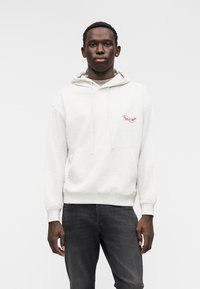 Ανοιχτό γκρι φούτερ με κουκούλα, μπροστινή τσέπη τύπου ποδιά, ρυθμιζόμενο κορδόνι και κόκκινο κεντημένο λογότυπο στο στήθος. Συνδυασμένο με σκούρο τζην.