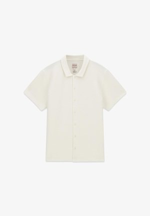 Polo manches courtes, blanc cassé, à boutons, col pointu, tissu texturé et étiquette de la marque Guess à l’intérieur du col.