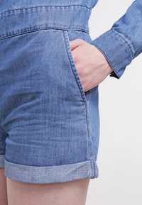 Jeansoverall i ljusblå färg, rullade ärmar och en sidoficka. Har en slät yta och synliga sömnadsdetaljer.