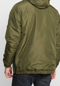 Olivengrøn polstret jakke med hætte, elastiske manchetter og en lynlåslomme på venstre ærme. Glat, blankt stof.