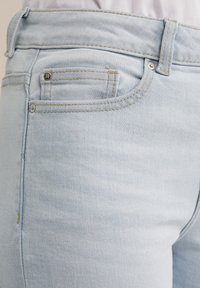 Jeans de denim azul claro con una textura suave, que cuentan con costuras en contraste, un pequeño bolsillo frontal y detalles metálicos en la cintura.