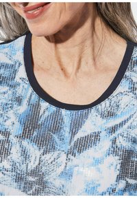 Korte mouwen top met een ronde halslijn, met een blauw en zwart pailletten bloemenpatroon op een lichte achtergrond en een donkere rand.