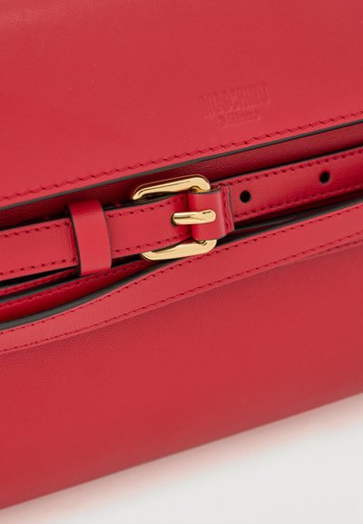 Κόκκινη δερμάτινη τσάντα με χρυσή αγκράφα και ρυθμιζόμενο λουράκι, με ανάγλυφο κείμενο του brand στο πάνω μέρος.