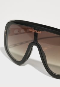 Occhiali da sole neri oversize con una montatura spessa, lenti marroni sfumate e un logo "CARRERA" dorato sul lato superiore destro.