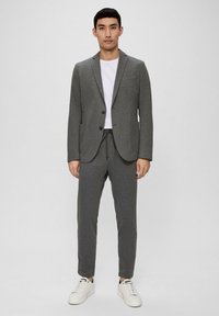 Grauer Blazer mit einem einzigen Knopf, seitlichen Taschen, kombiniert mit passenden Jogginghosen aus weichem Material, abgerundet mit weißen Turnschuhen.