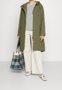Oliwkowa parka z kapturem, z metalowymi detalami w kolorze srebrnym oraz zapięciem na guziki. Połączona z paskowanym topem, szerokimi białymi spodniami oraz w kratkę torbą.