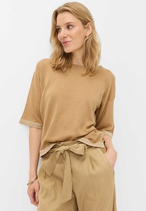 Blonde vrouw draagt een beige trui met korte mouwen en beige high-waisted broek met een strik, staand met één hand in de zak.