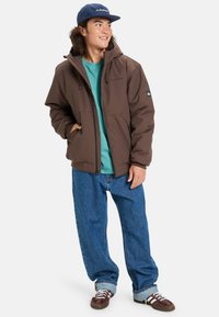Ung man i brun huva jacka, teal t-shirt, lösa blå jeans, vinröda sneakers och en marinblå keps, står med händerna i fickorna.