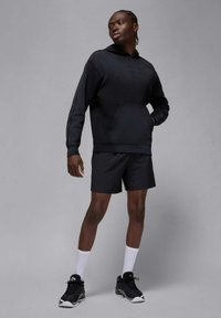 Czarna bluza z kapturem z przednią kieszenią i sznurkiem, zestawiona z czarnymi spodenkami. Do tego sportowe buty i białe skarpetki za kostkę. Neutralne szare tło.